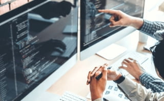 プログラミングのコードが映ったPCディスプレイを見ながら話し合う2人の写真